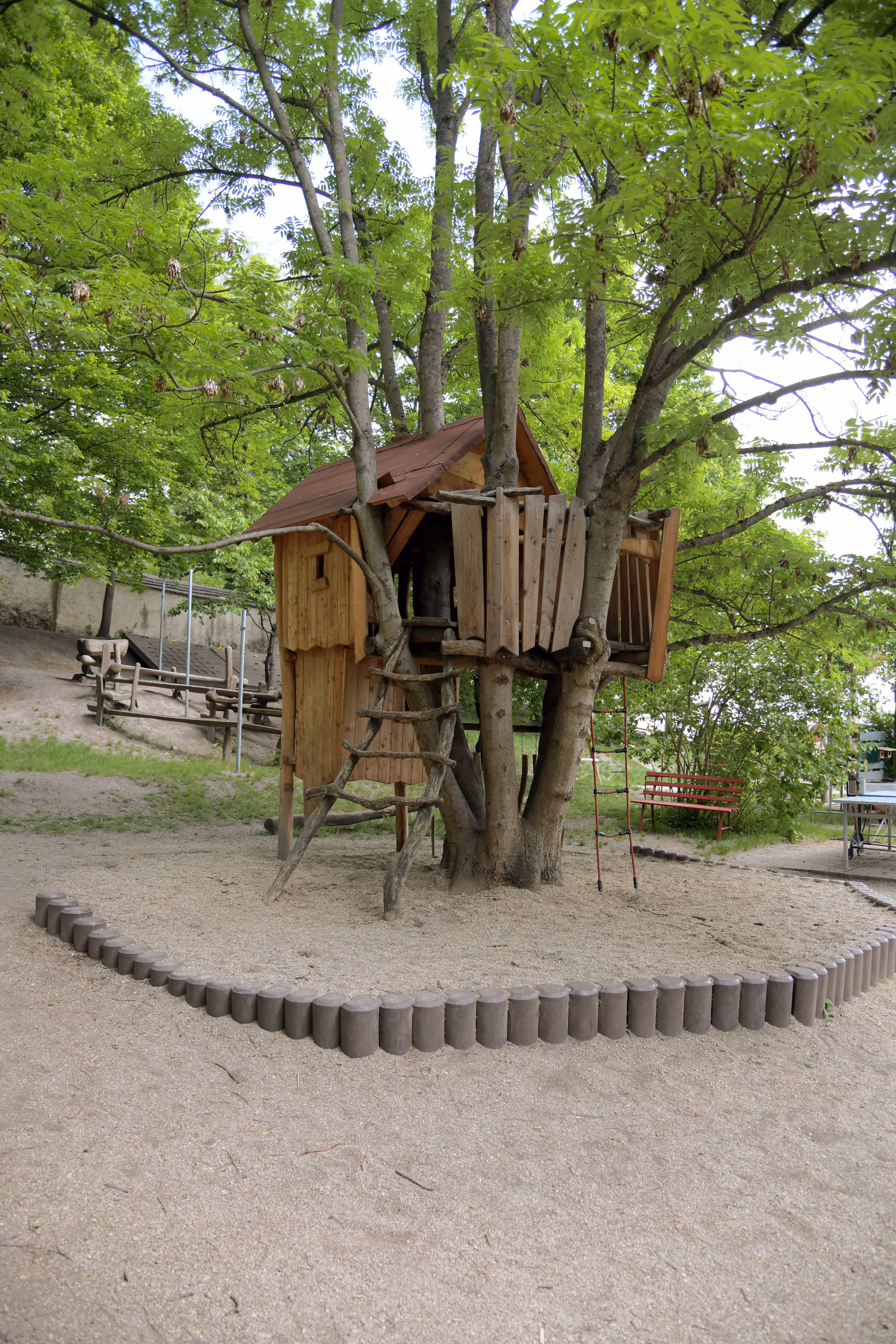 Baumhaus zwischen Bäumen mit Sandbereich davor.