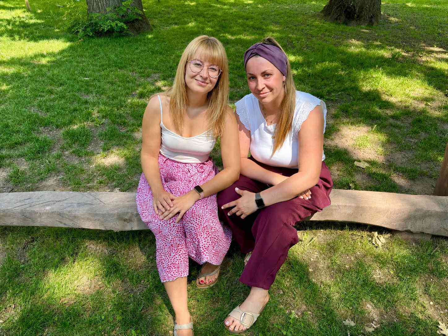 Zwei Frauen sitzen lächelnd auf einem Baumstamm im Grünen.