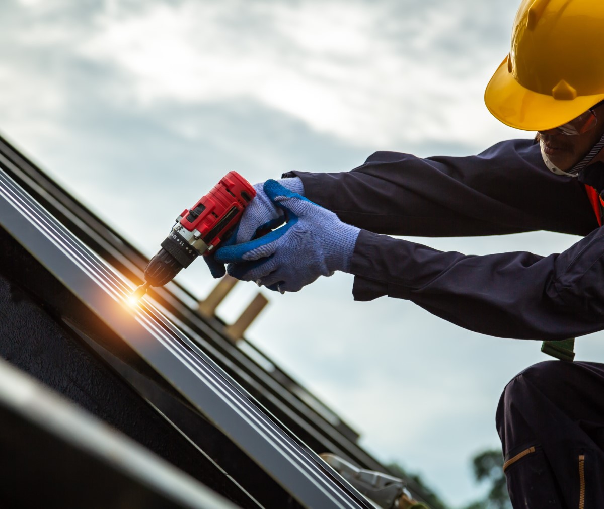 Construction worker in yellow helmet using power drill on metal roof