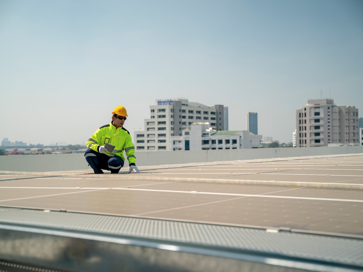 Technician in safety gear inspecting solar panels on a rooftop with city buildings in the background.
