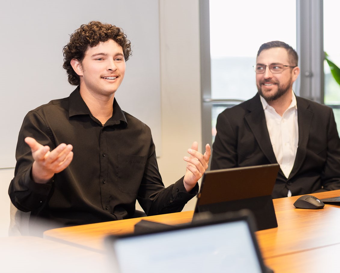 Two men in business attire engaged in a discussion at a meeting room table.
