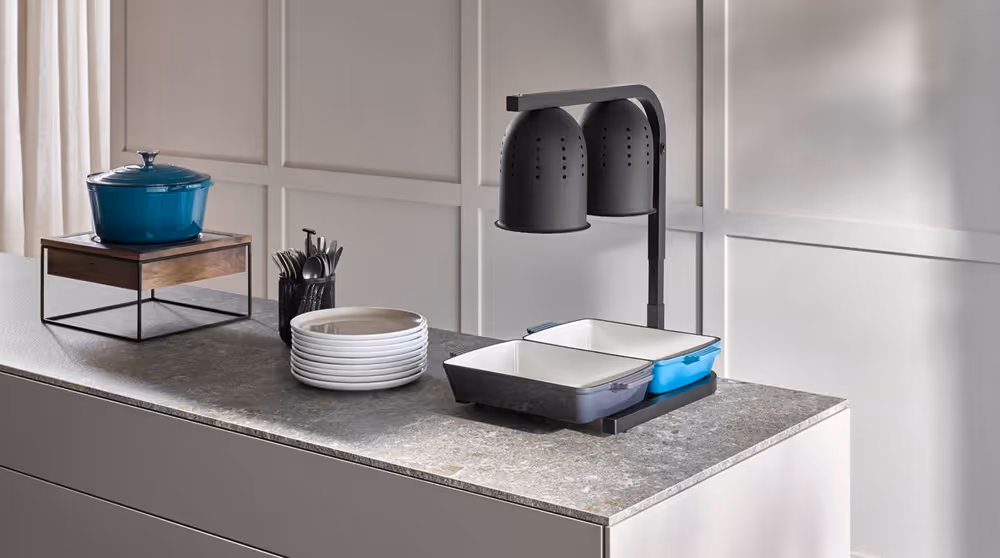 Buffetstation mit Wärmelampen, Servierbehältern, Tellern und Besteck auf einer modernen Arbeitsfläche.