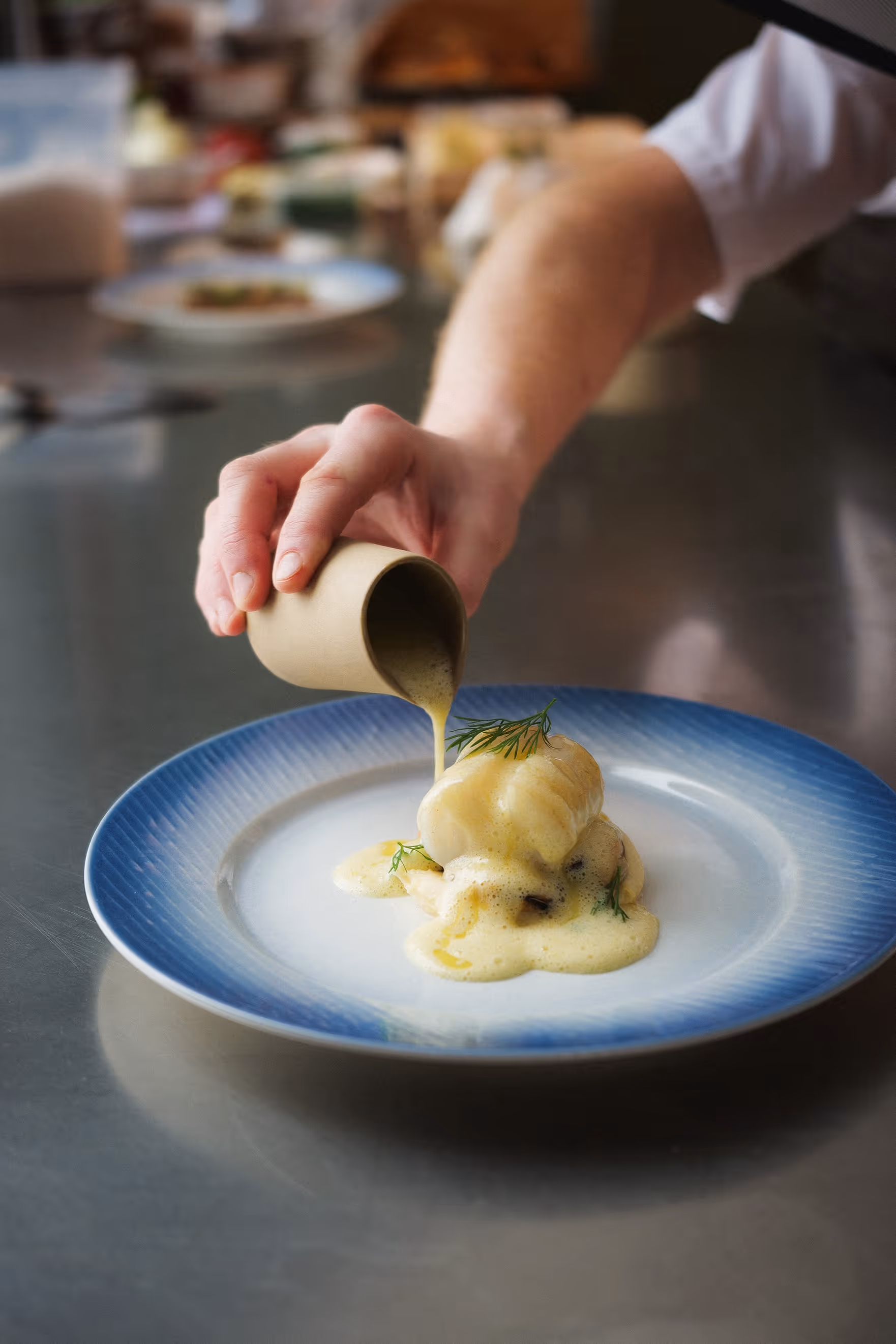 Koch richtet ein Gericht an und giesst eine Sauce auf einem blauen Porzellanteller an.