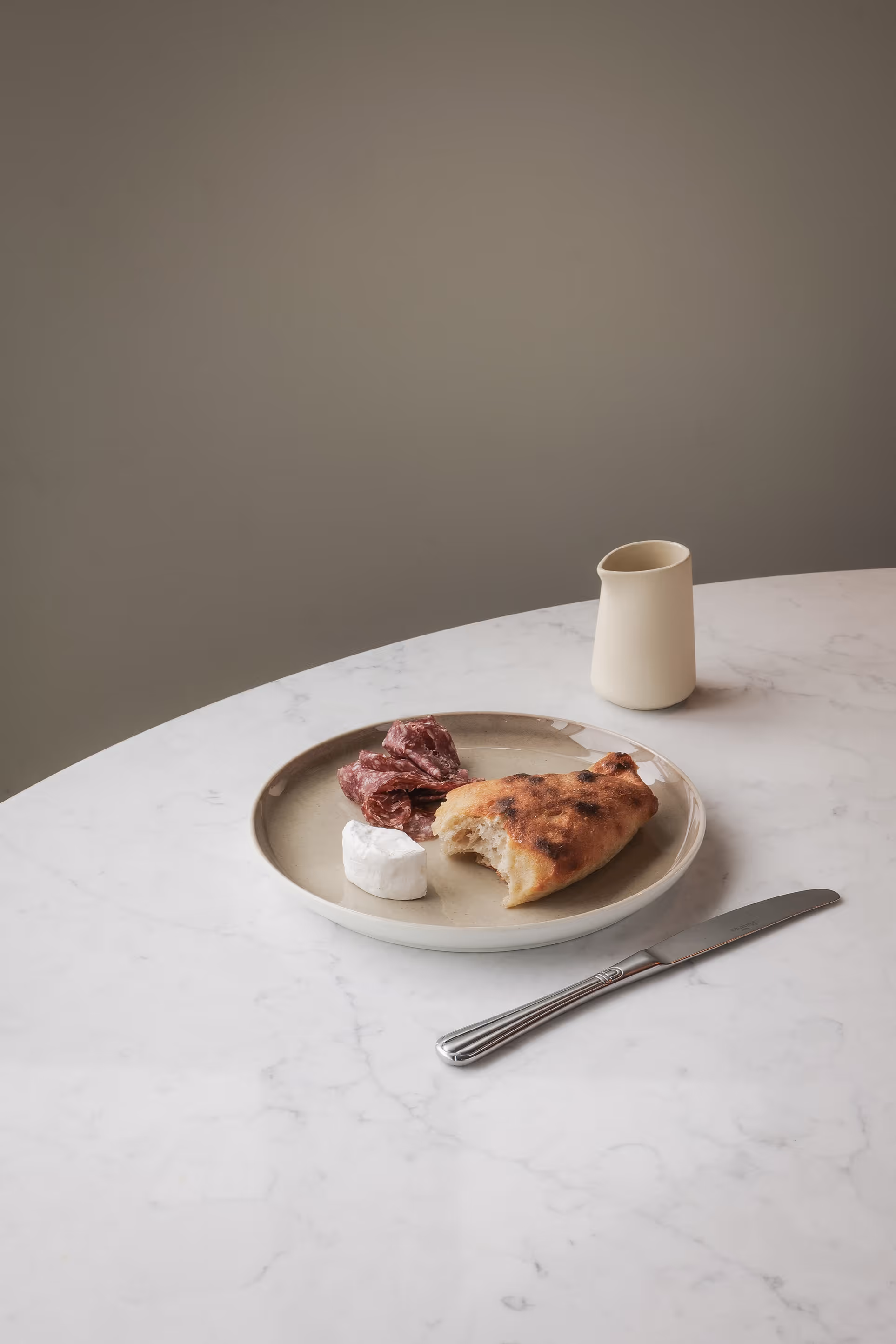 Keramikteller mit Brot, Käse und Salami auf einem Marmortisch in schlichtem, modernem Ambiente.