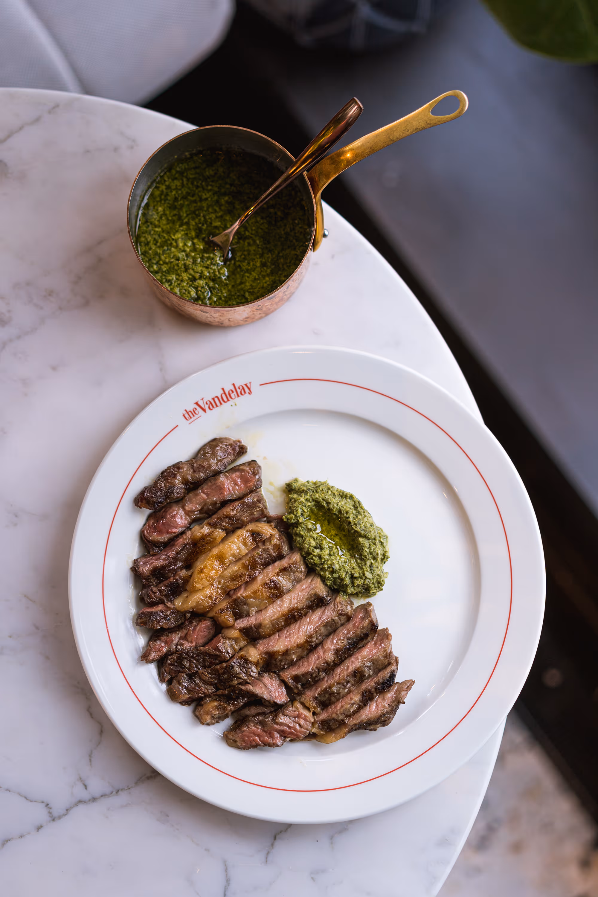 Geschnittenes Steak auf einem Porzellanteller mit grüner Kräutersauce und Kupfertopf.