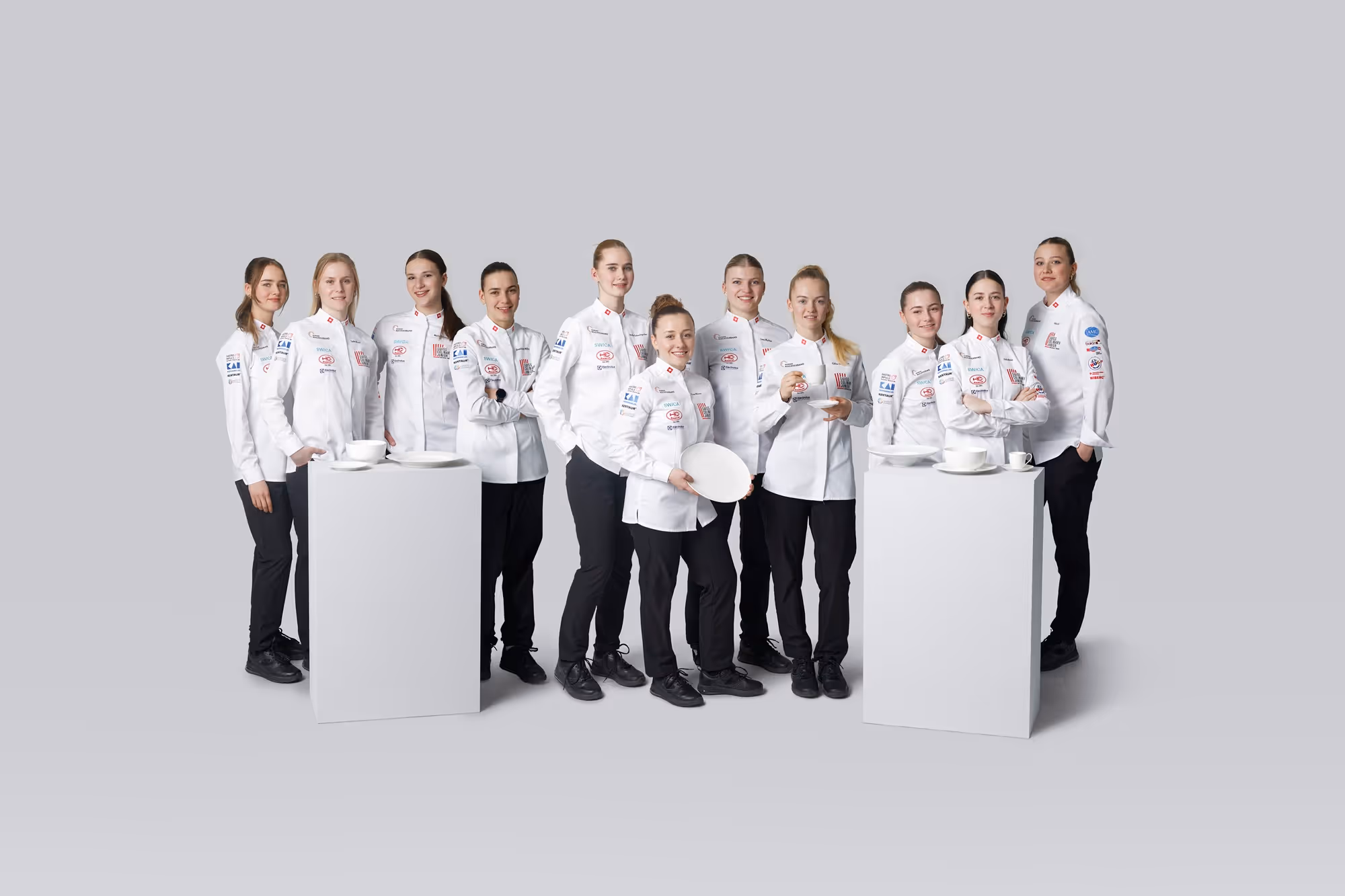 Mitglieder der Junioren Kochnationalmannschaft posieren in ihrer offiziellen Kochbekleidung für ein Gruppenfoto mit Geschirr und Tassen.