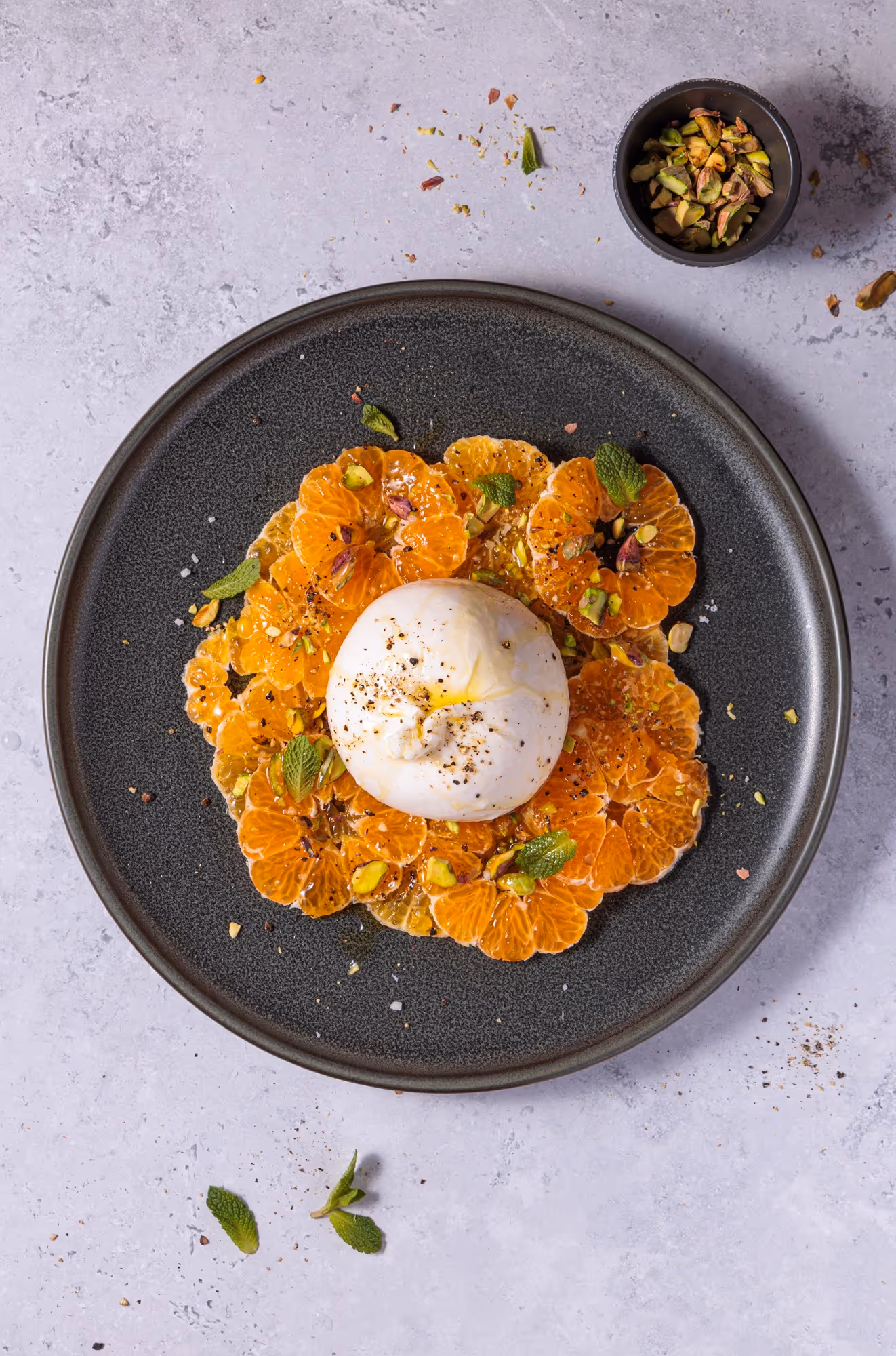 Dessert mit Burrata, Mandarinen und Pistazien auf einem dunklen Teller, serviert mit Besteck.