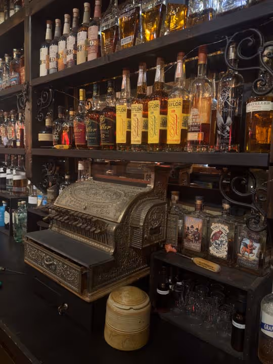 Shelves with various liquor bottles and an antique ornate cash register in a bar setting.