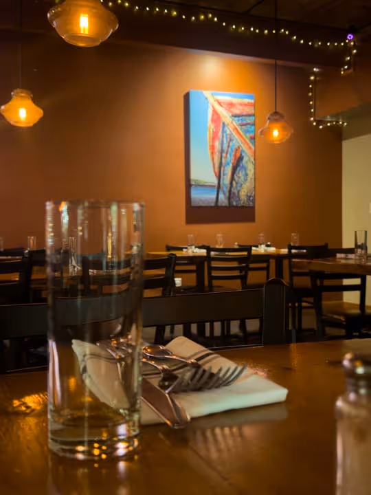 Interior of a cozy restaurant with wooden tables set with utensils and glasses, warm hanging lights, and a colorful boat painting on an orange wall.