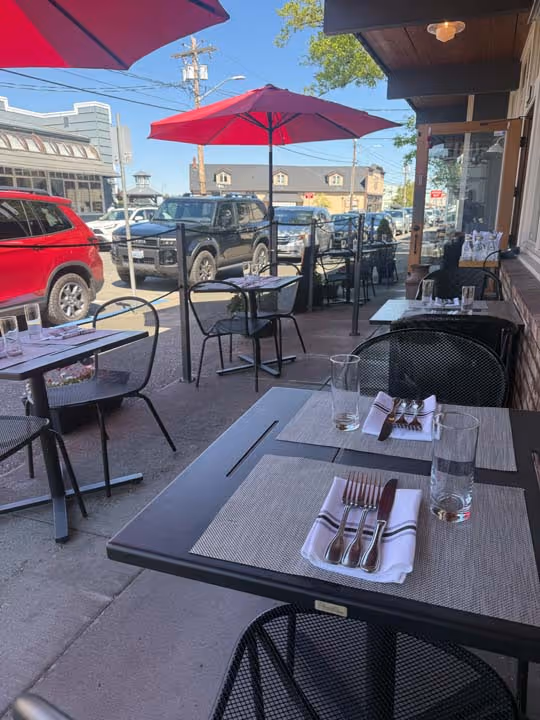 Outdoor restaurant seating with black metal tables and chairs, place settings, red umbrellas, and parked cars along the street in the background.