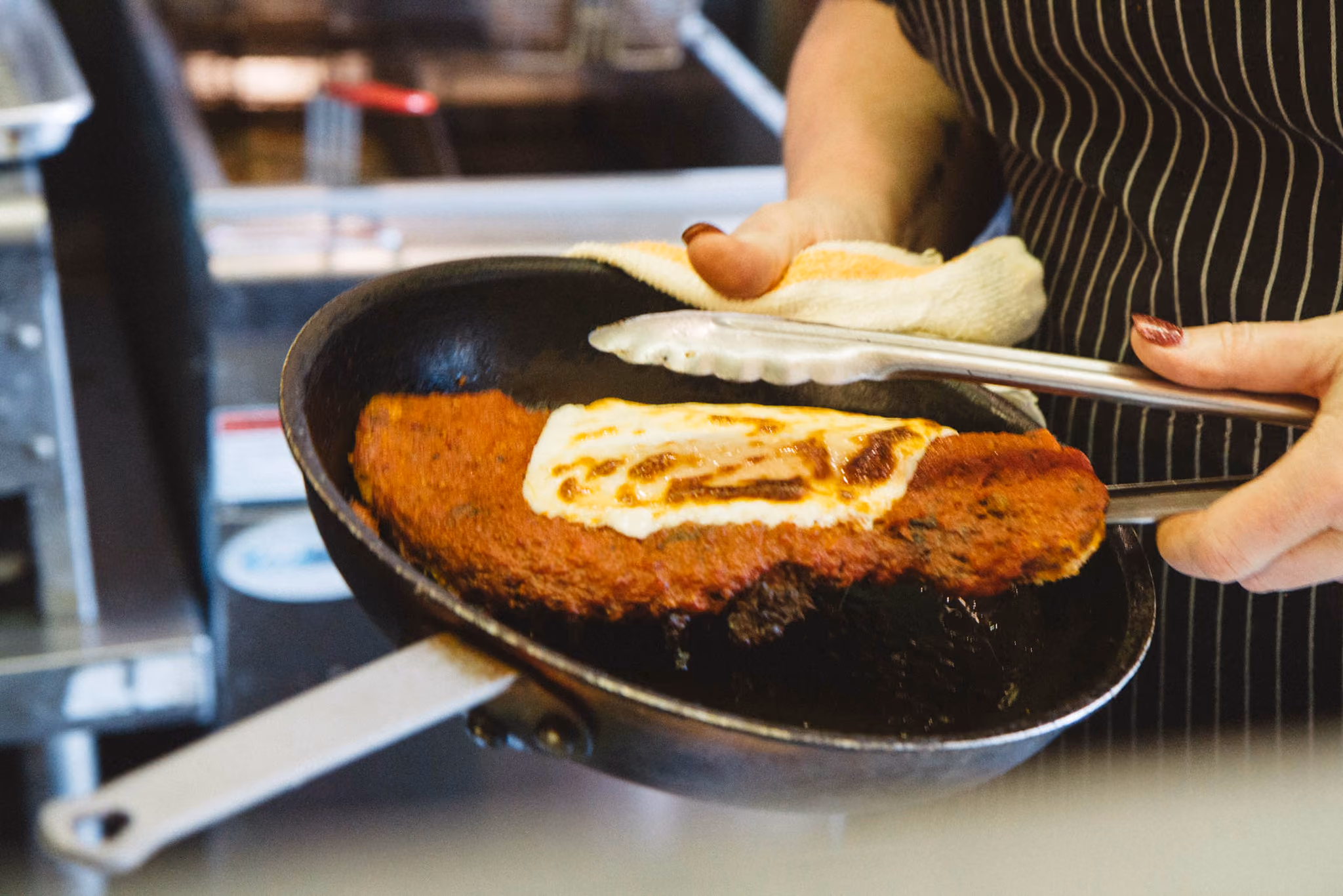 Person holding a frying pan with cardboard and metal tongs grilling halloumi cheese on a breaded vegan schnitzel patty in a kitchen.