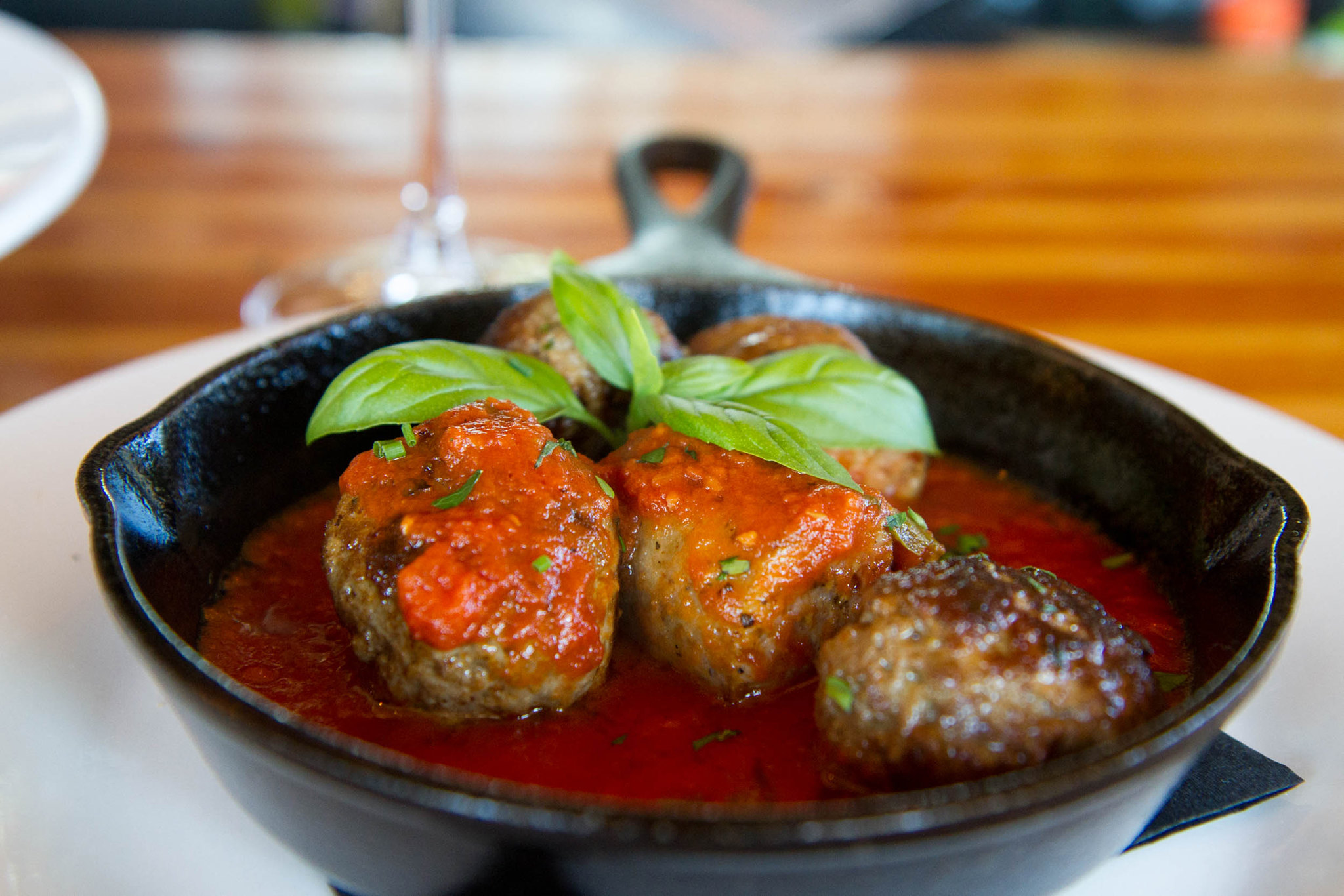 Cast iron skillet with several meatballs topped with tomato sauce and fresh basil leaves, placed on a white plate on a wooden table.