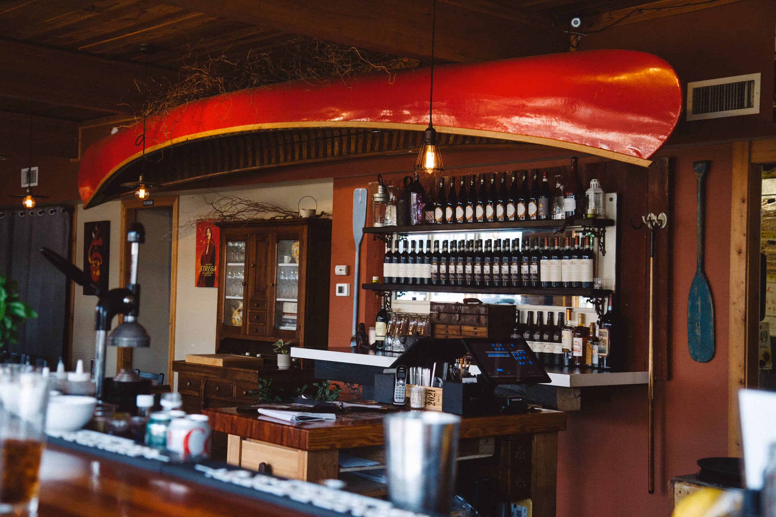 Cozy bar interior with a red canoe mounted above the counter, shelves filled with bottles, a wooden paddle hanging on the wall, and warm hanging light bulbs.