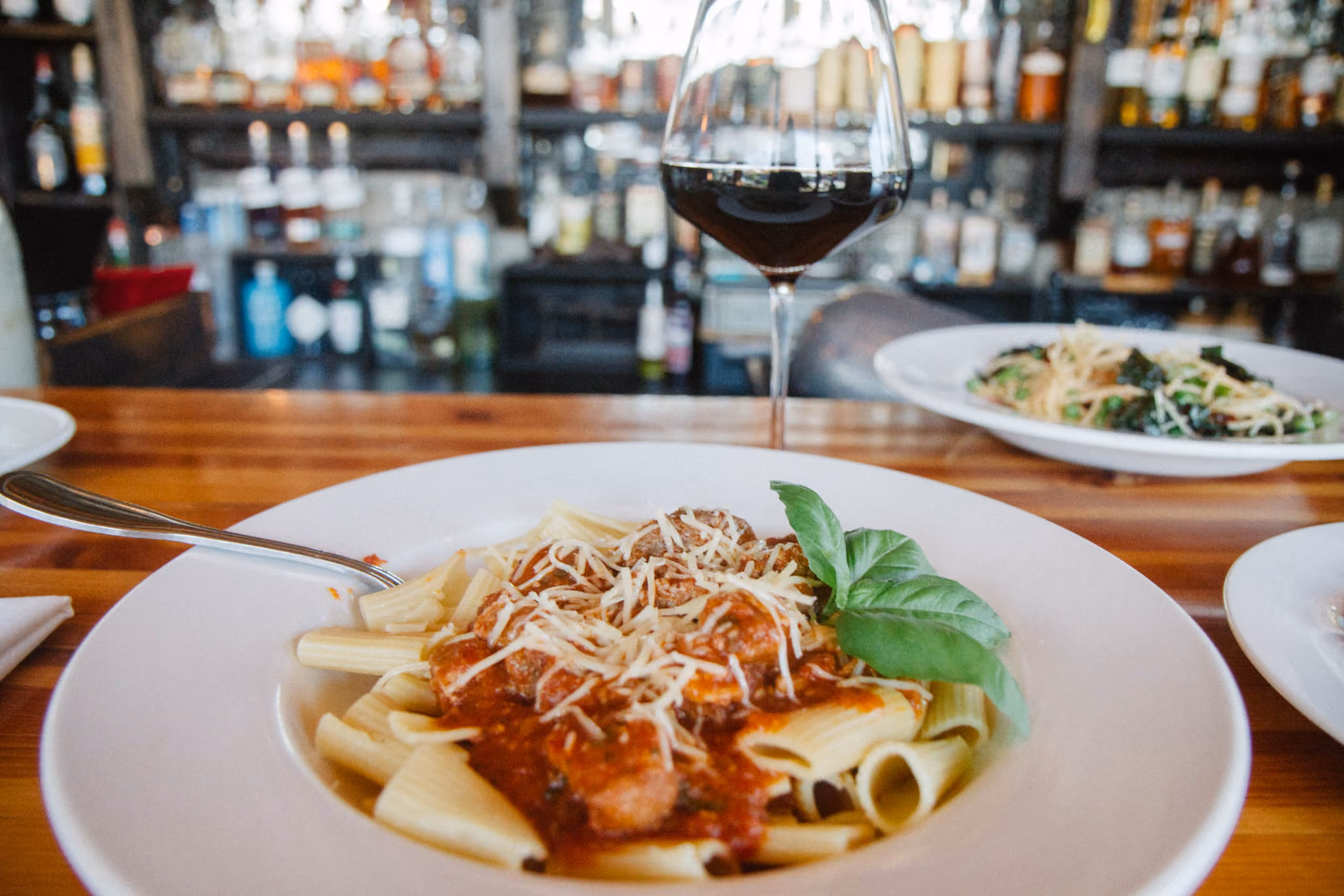 Plate of rigatoni pasta topped with tomato sauce, meatballs, grated cheese, and a basil leaf on a wooden bar counter with a glass of red wine and bottles in the background.