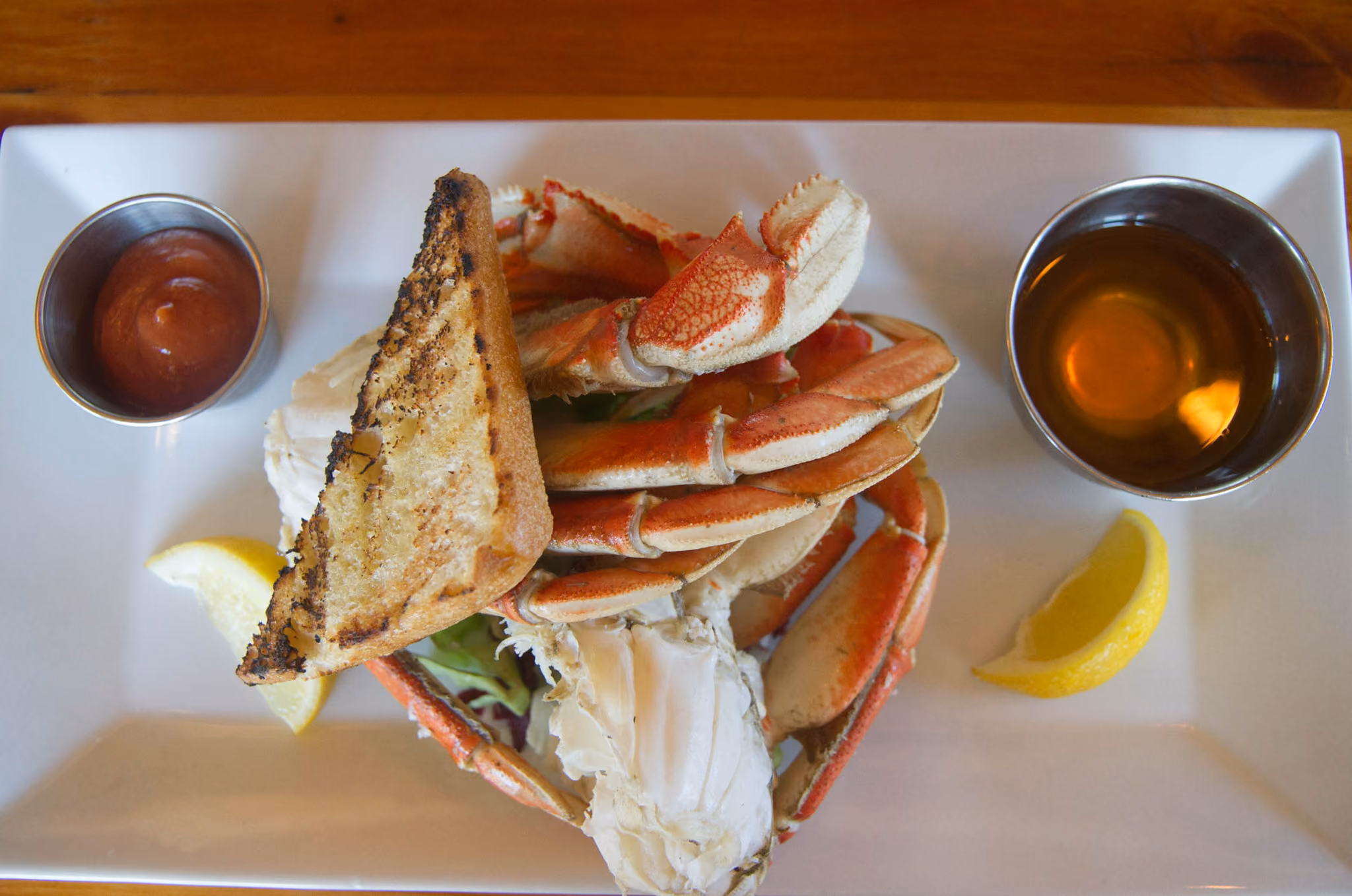 Plate with cooked crab legs, two lemon wedges, a piece of grilled bread, a small metal cup of cocktail sauce, and a small metal cup of melted butter.