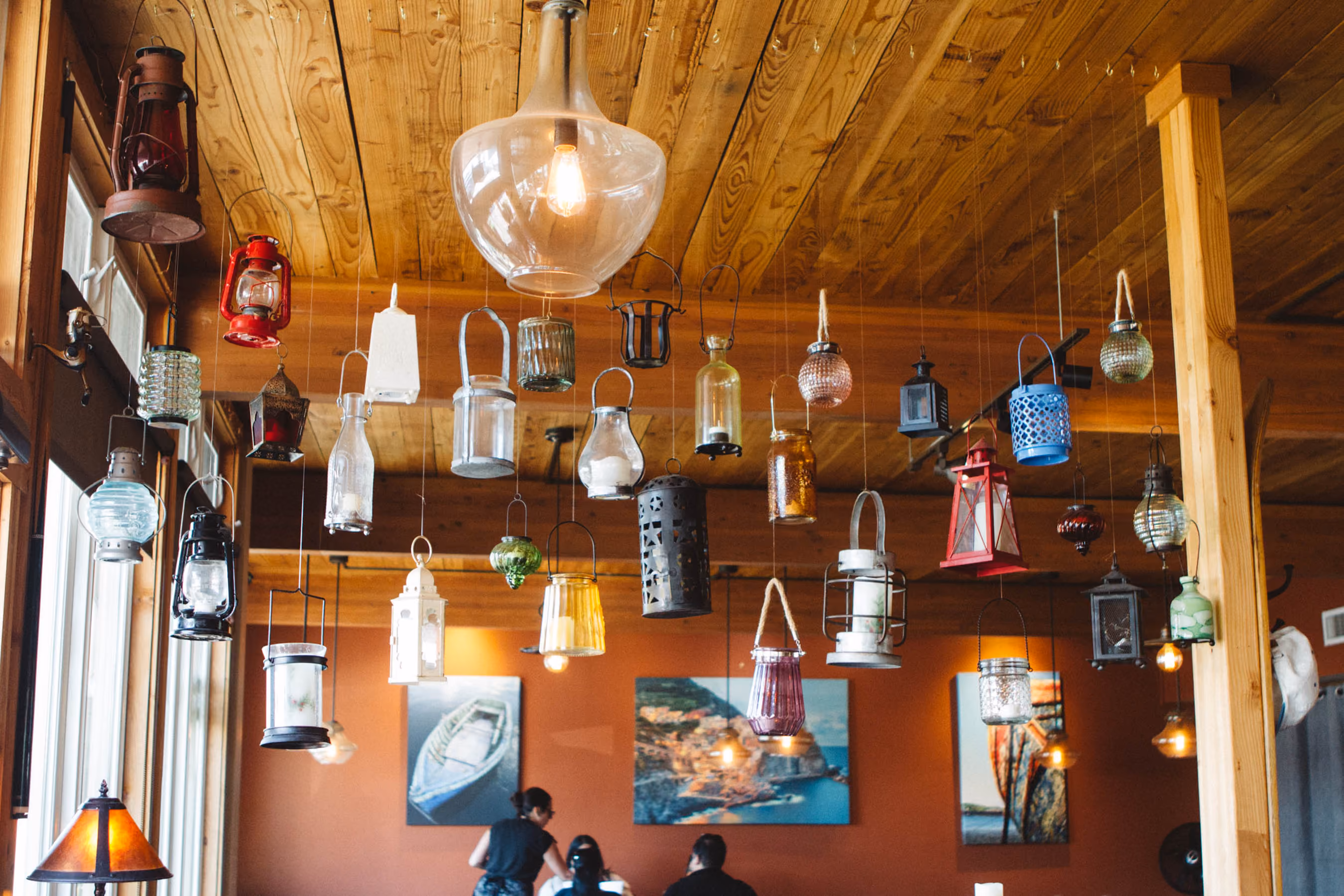 Interior of a room with a wooden ceiling decorated with numerous hanging vintage lanterns of various colors, shapes, and sizes, with a large clear glass pendant light in the center and three people sitting at a table below near wall art.