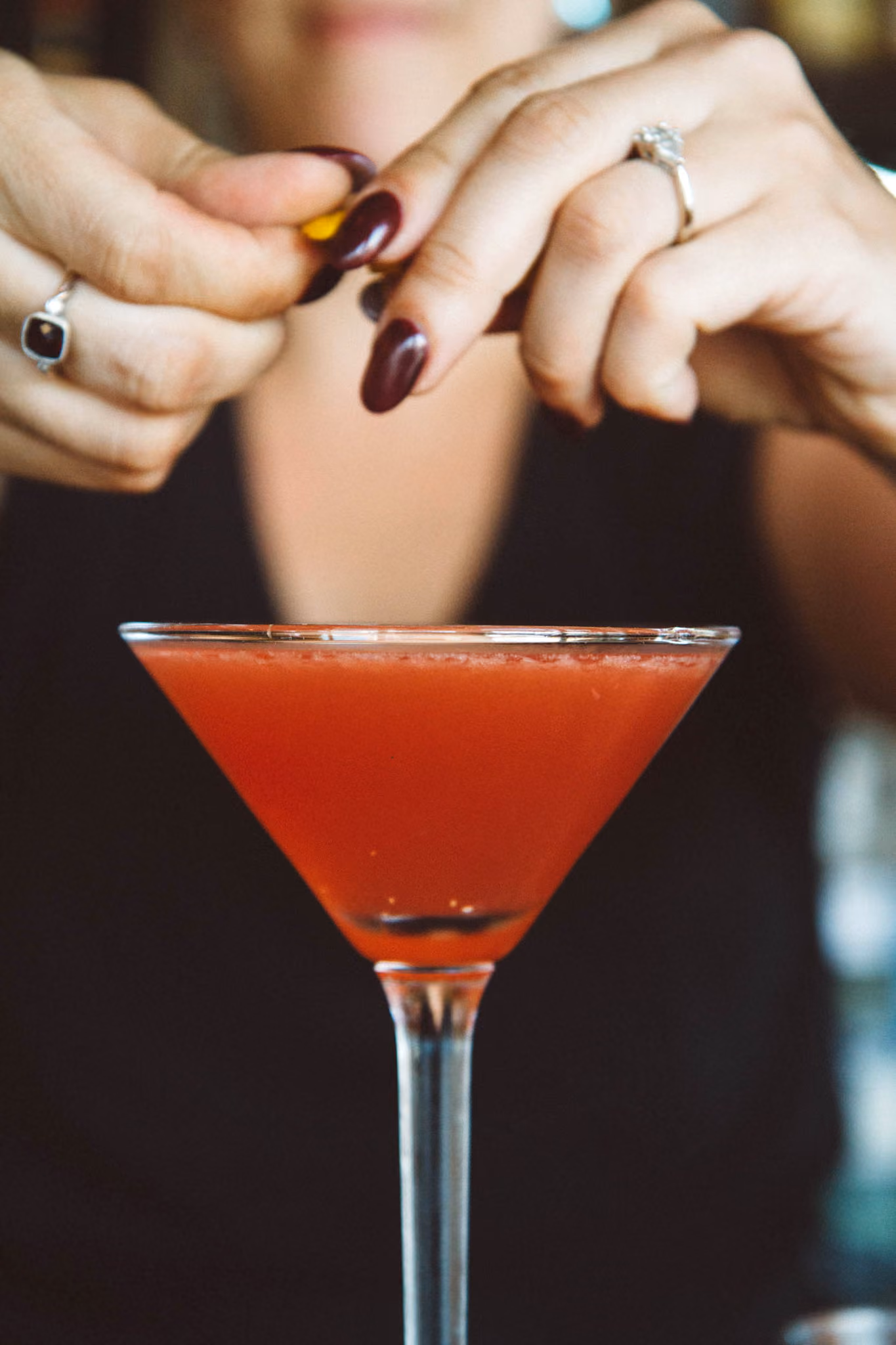 Close-up of a person with dark nail polish and rings garnishing a red cocktail in a martini glass with a citrus peel.