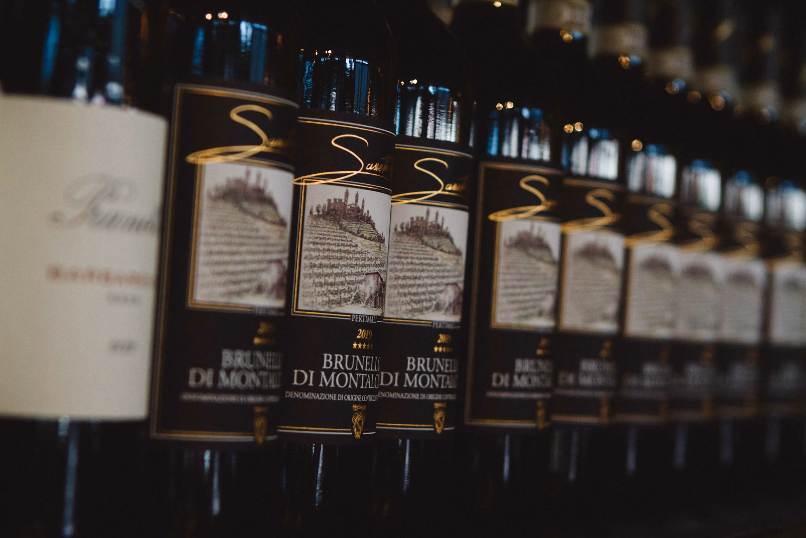 Rows of Brunello di Montalcino red wine bottles with black and gold labels lined up on a shelf, focused on the front bottles while the background bottles are blurred.