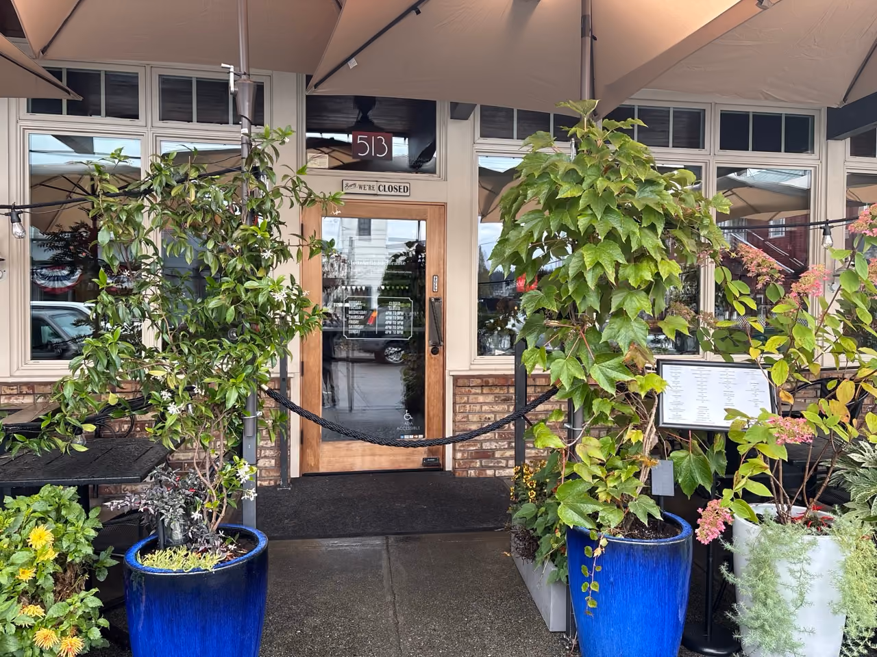 Entrance of a closed establishment with a wooden door marked '513' and 'We’re Closed' sign, flanked by large blue pots containing leafy green plants under a canopy.