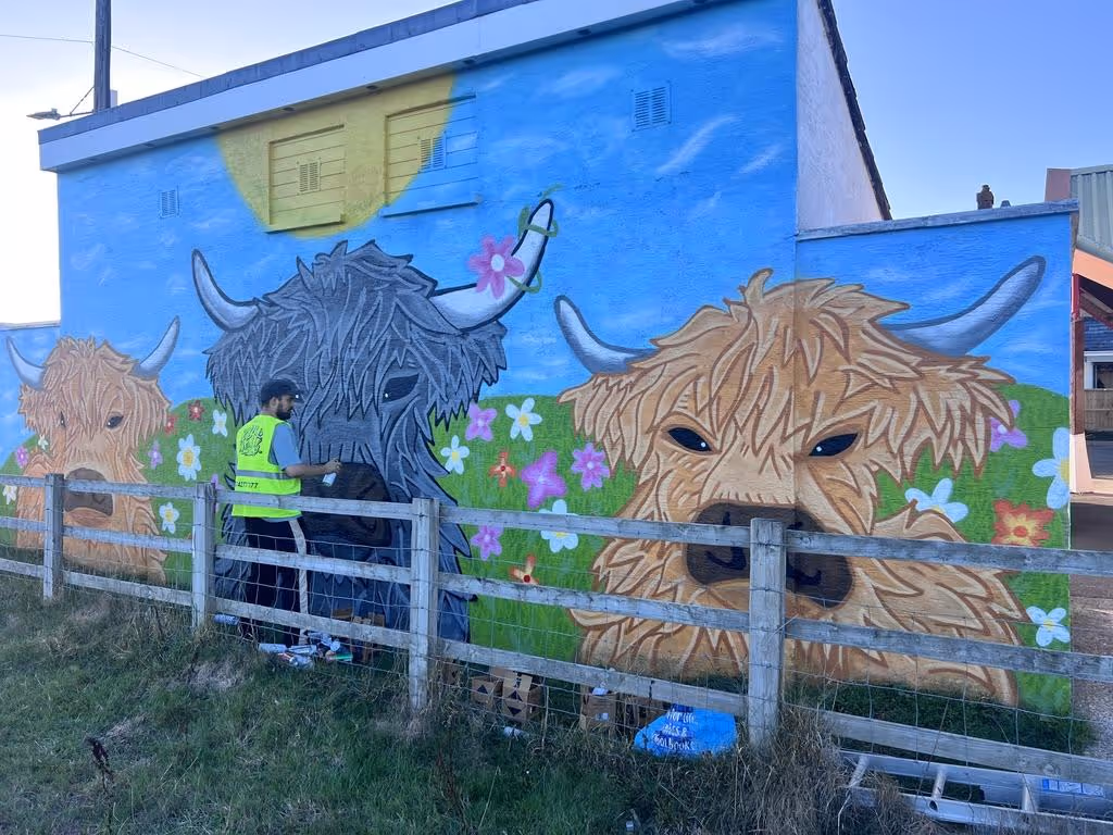Artist painting a colorful mural of three Highland cows with flowers on a blue sky and green grass background on a building wall.
