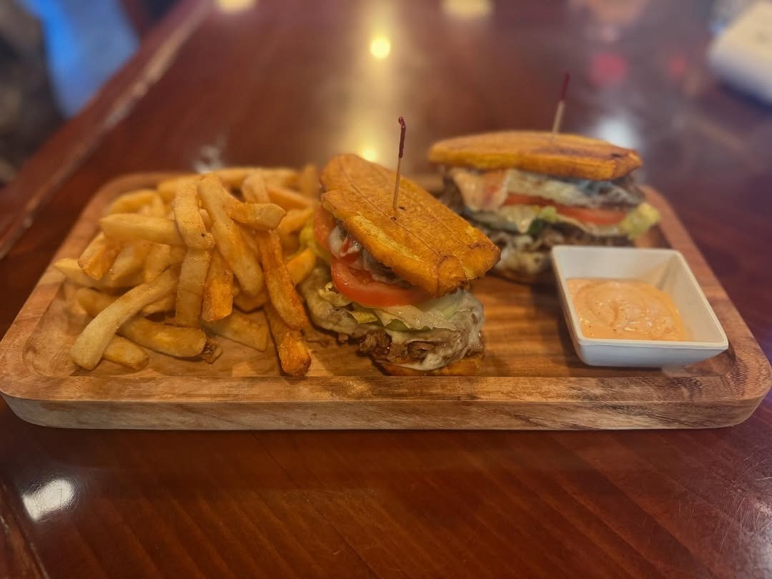 Wooden platter with two sandwiches made of plantain buns, lettuce, tomato, cheese, and meat, served with French fries and a side of dipping sauce.