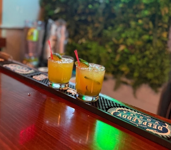 Two yellow-orange cocktails with ice, red straws, and lime wedges served on a bar counter with a Deedee Vodka mat.