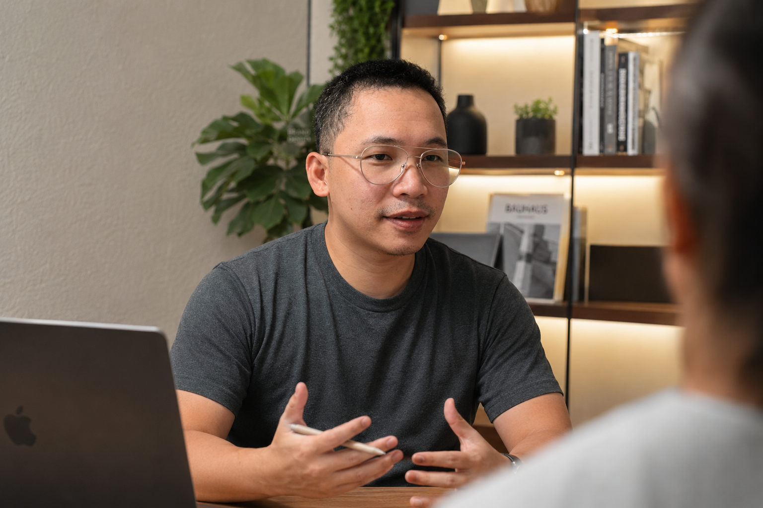 Man in glasses talks with another person across a table with a laptop in a modern office.