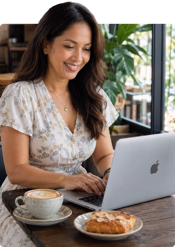 Smiling woman using a laptop at a cafe table with coffee and pastry nearby.