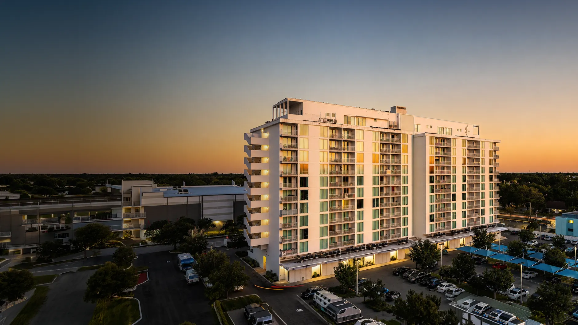 Modern multi-story apartment building with balconies at sunset and cars in a parking lot below.