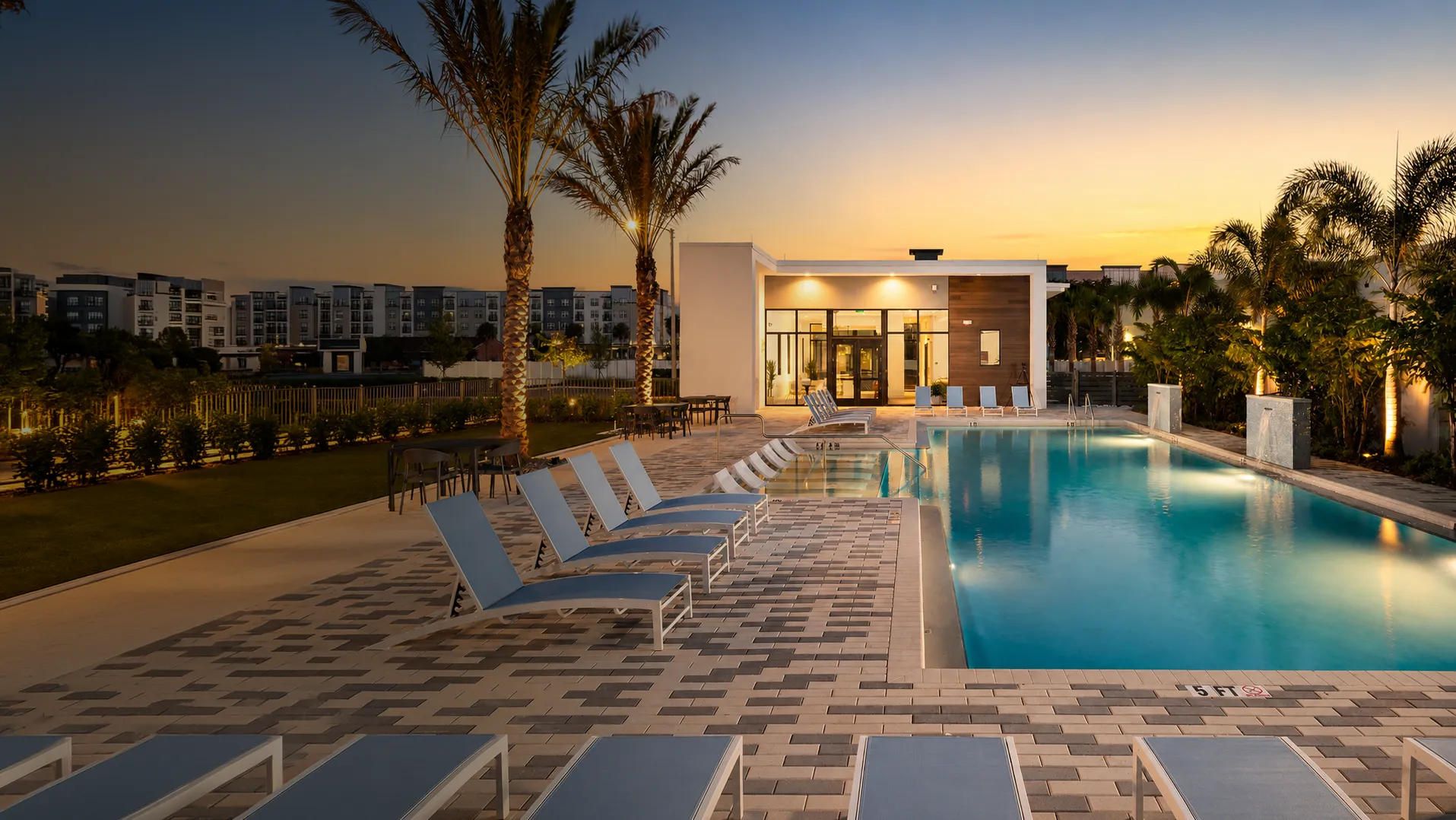 Modern outdoor swimming pool with lounge chairs and palm trees at sunset near apartment complex.