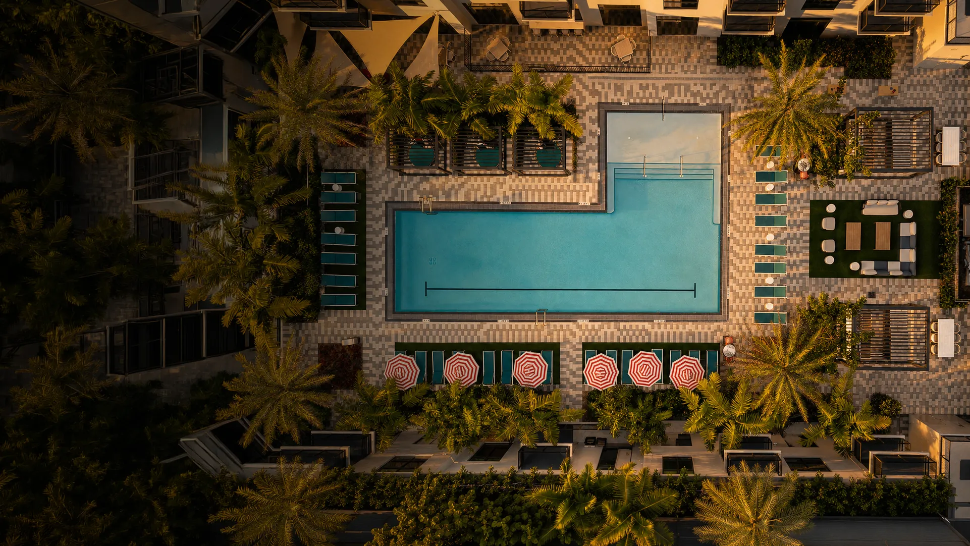 Aerial view of a rectangular swimming pool surrounded by palm trees and lounge chairs with red umbrellas.