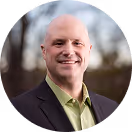 Smiling bald man in a black blazer and green shirt outdoors with blurred trees background.