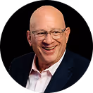Smiling bald man with glasses wearing a dark suit jacket and white shirt against black background.