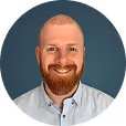 Smiling man with red beard and shaved head wearing a light blue collared shirt on blue background.
