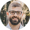 Smiling man with beard, glasses, and short hair outdoors with blurred greenery background.