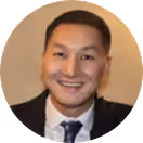 Smiling man wearing a black blazer, white shirt, and black tie with a blurred beige background.