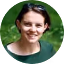Woman with short dark hair smiling outdoors with green foliage in the background.