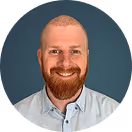 Smiling man with red beard and mustache wearing a light blue button-up shirt against a solid background.