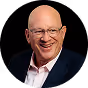 Bald man wearing glasses and a dark blazer smiling against a black background.