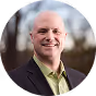 Smiling bald man wearing a light green shirt and dark blazer outdoors with blurred background.