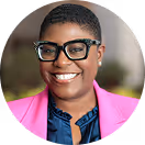 Smiling person wearing black glasses, a pink blazer, and a blue shirt with short hair.