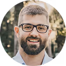Smiling man with glasses and beard, outdoors with blurred natural background.