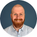 Smiling bald man with red beard wearing a light blue button-up shirt on a teal background.