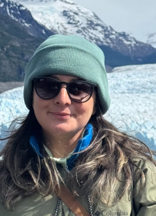 Woman wearing sunglasses and green beanie with snow-covered mountain and glacier in the background.