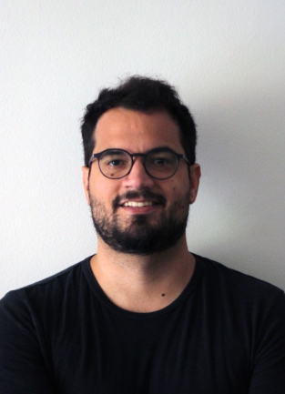 Man with dark hair and beard wearing glasses and a black shirt smiling in front of a light wall.