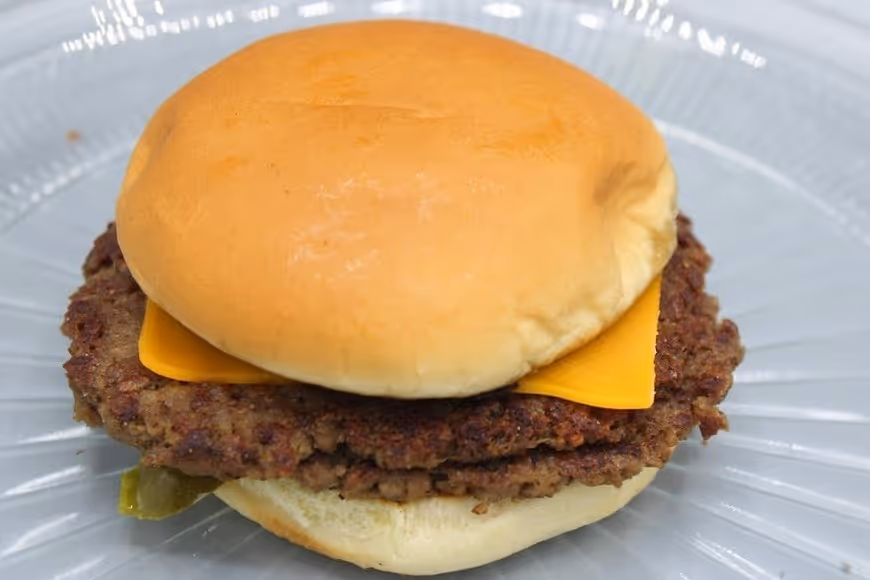 Cheeseburger with two beef patties, a slice of cheddar cheese, and a soft bun on a clear glass plate.