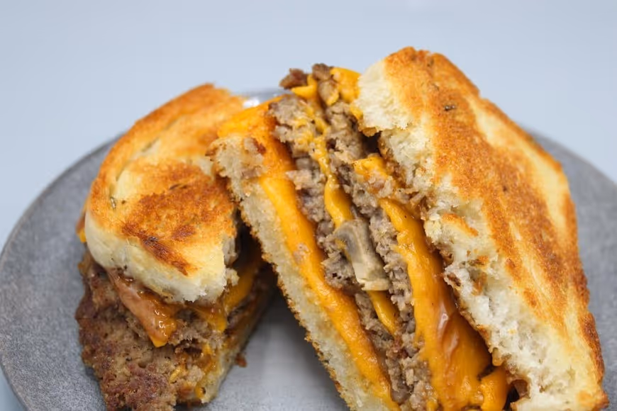 Grilled sandwich with melted cheddar cheese, seasoned ground beef, and mushrooms on toasted bread halves on a gray plate.