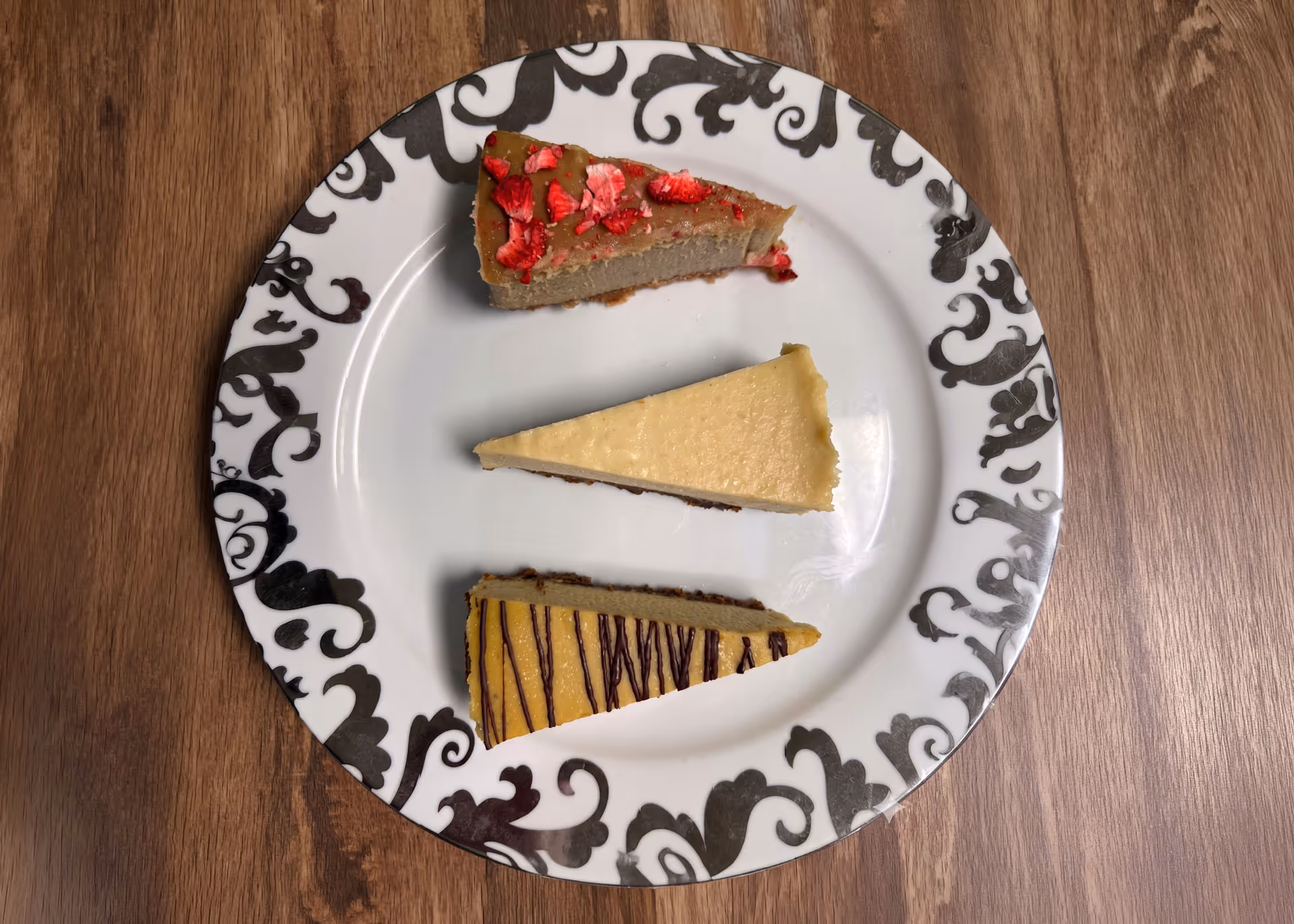 Three slices of cheesecake on a white plate with black decorative patterns, placed on a wooden surface.