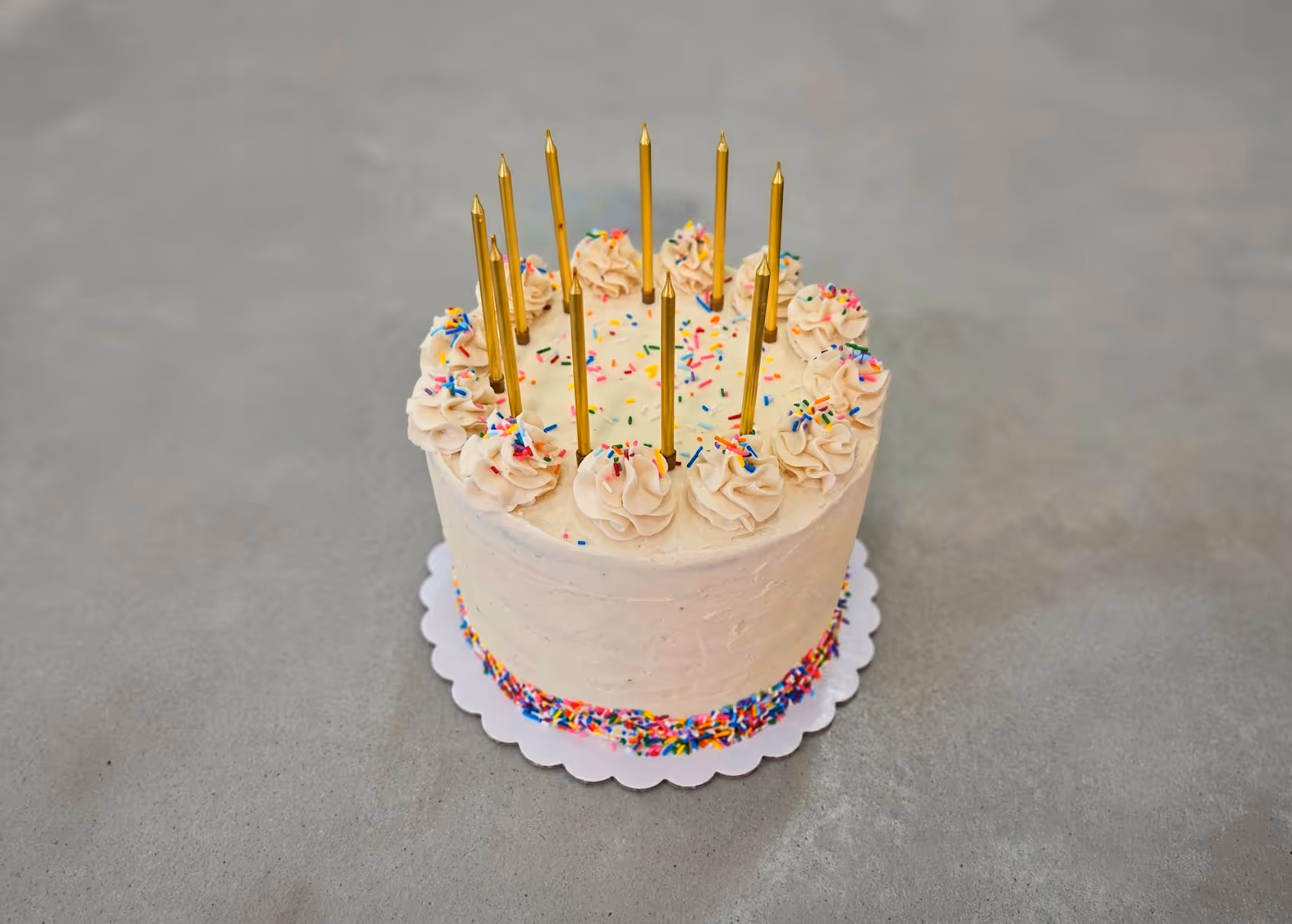 A round vanilla cake with cream frosting, colorful sprinkles, and tall gold candles.