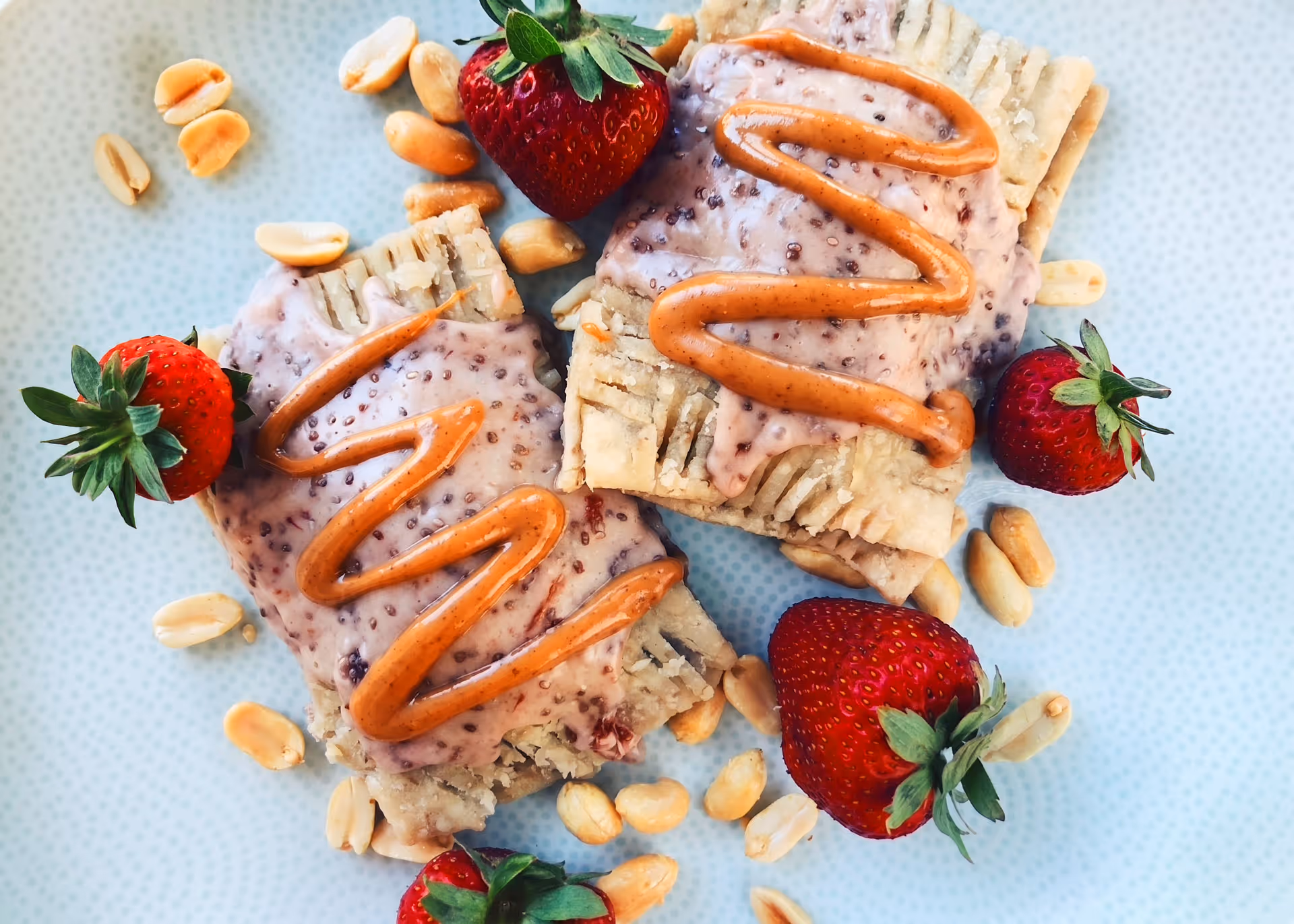 Two pastry squares topped with pink cream and peanut butter drizzle, surrounded by fresh strawberries and peanuts on a light blue plate.